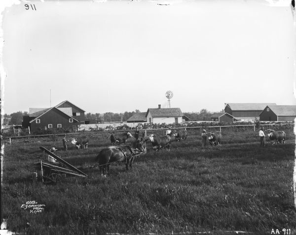 Farm house, barn, and farm buildings surround by fence.  Trees and windmills in background. Man riding horse-drawn corn binder in field with many men and cattle.