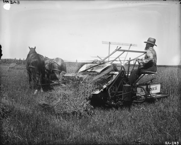 Side view of a bearded man wearing a braided hat operating a McCormick grain binder in a field.