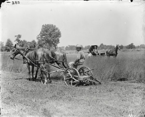 View towards a man wearing a banded hat operating a horse-drawn mower in a field. In the background there is a boy standing near a man in a horse-drawn carriage.