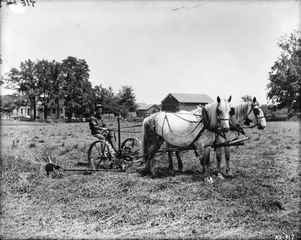 Man operating a horse-drawn mower in a field. In the background is a farmhouse and other buildings surrounded by trees and a fence.
