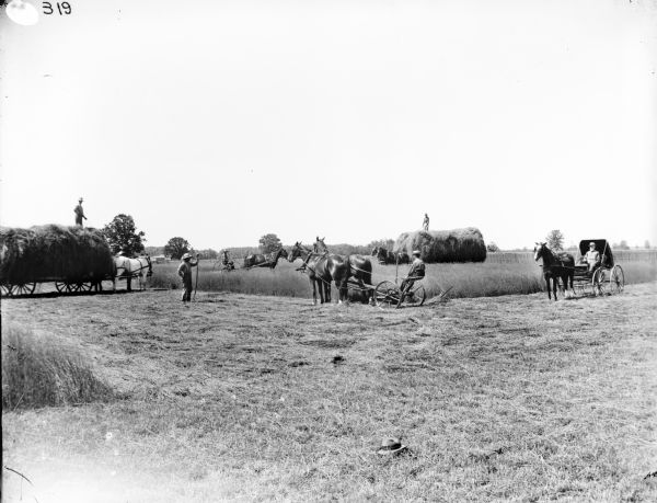 Men working on haying operation in a field. Two men are driving horse-drawn wagons piled high with hay. One man is standing near one wagon holding an implement, and on the right a bearded man is driving a horse-drawn carriage. In the foreground a hat is lying in the field.