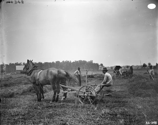 Men with a horse-drawn mower in a field. One man is working with a hand implement in a pile of hay, and another is sitting in a horse-drawn carriage in the background.