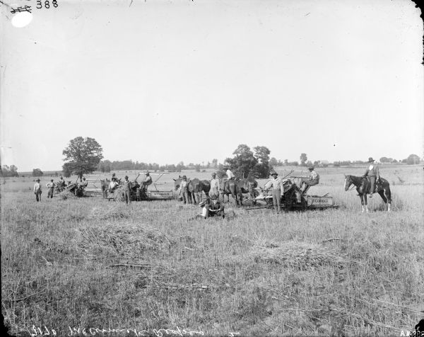 A large group of men, mostly African American, posing in a field with three McCormick grain binders. Each binder is being operated by a man and is pulled by two horses. There is McCormick signage on the binders. One man is posing on the right sitting on a horse, and two men are sitting in the field in the foreground. Farm buildings are in the background.