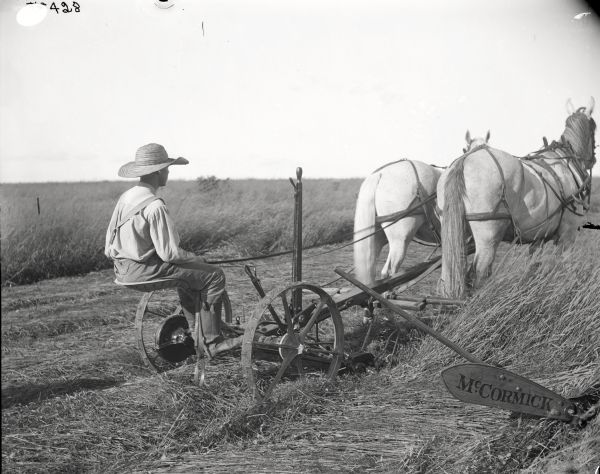 Rear view towards right side of a young man, wearing a wide brimmed hat, operating a McCormick mower pulled by a team of two horses.
