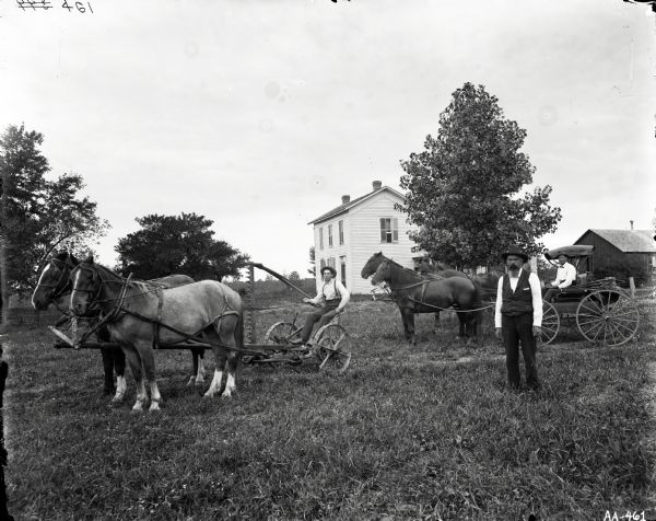 Three men are posing in front of a farmhouse with farm equipment and carriages. One man is standing in the field, another man is driving a mower, and a third man is sitting in a carriage pulled by two horses. Two other horses are directly behind, perhaps pulling another carriage.