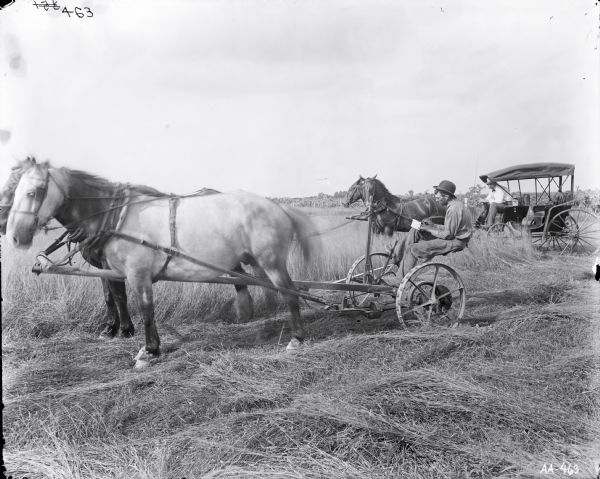 Side view of a man sitting on a horse-drawn mower in a field. The man on the mower is writing on a piece of paper. Another man wearing a bow tie is sitting on a horse-drawn carriage behind him.