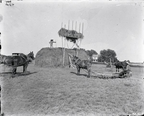 A group of men are working in a field with farm buildings in the background. A man wearing a wide brimmed hat is standing on top of a large pile of hay holding a farm implement. Another man is using a wooden horse-powered hay stacker to lift hay to the top of the pile.