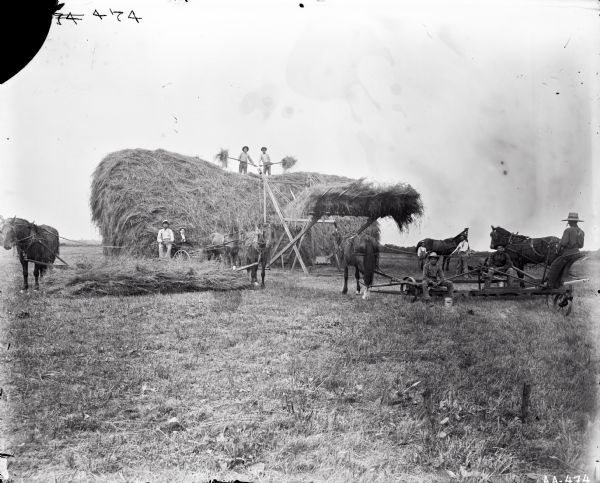 Outdoor group portrait of men harvesting hay. Two men are standing on top of a haystack with pitchforks filled with hay. A wooden counter-weighted hay stacker is in the process of lifting another pile of hay to the top of the pile. Several men are posing at the base of pile. Two horses are dragging a pile of hay between then. Several other men and horses are standing on the right.