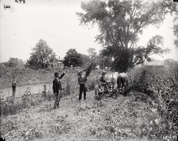 One man is operating a horse-drawn mower in a field. Two men are standing nearby, with one man pointing up toward the sky, and the other man holding a bunch of plants. Behind them is a fence and a dirt road. A farmhouse and trees are in the background.