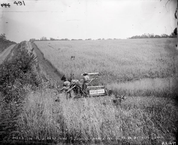 View down hill of a man in wide brimmed hat operating a horse-drawn McCormick grain binder in a field. A fence and road run parallel to the field on the left. Text written on glass plate is as follows: [MT? Work on A.O. Watsons 400 acre farm 5 miles N.W. Albia Iowa.]