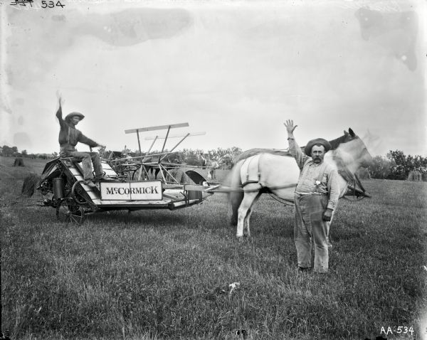 Group portrait of men and boys waving in a field. One man is operating a horse-drawn McCormick grain binder. Another man and a boy are in a horse-drawn buggy behind him. In the middle a young man is standing with a dog, and behind him is a man waving with a hat. Another man is waving in the right foreground.