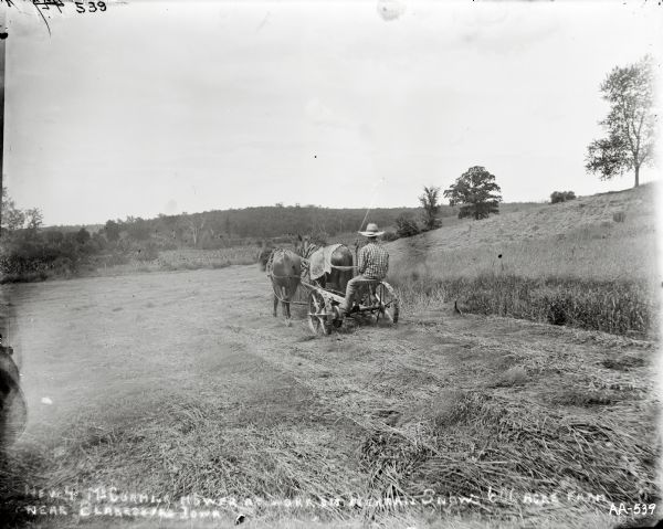 View from behind of a man with a wide-brimmed hat is operating a horse-drawn mower near trees and a low hill. Text written on the glass place is as follows: [New 4 McCormick Mower At Work on Herman Snows 600 Acre Farm Near Blakesburg Iowa].