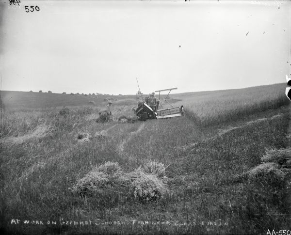 View from behind of a man operating a horse-drawn McCormick grain binder in a field. Another man with a beard is standing nearby. Text written on glass plate is as follows: [At work on Gephart Schoegh's Farm near Blakesburg, IA]