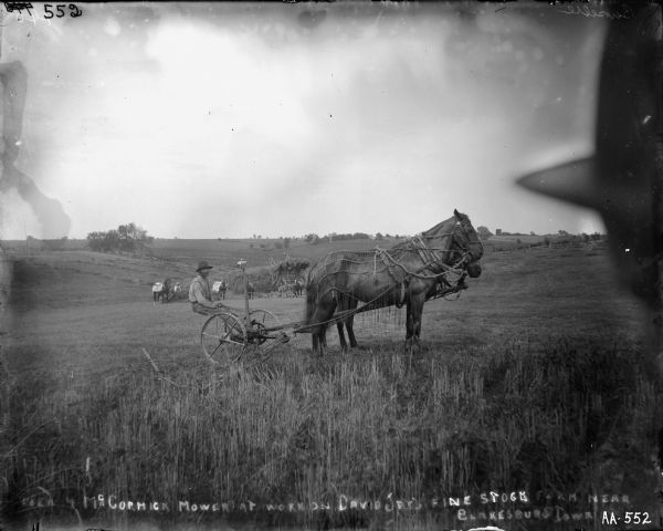A man is posing on a horse-drawn mower in a field. Behind him a group of men are stacking hay using a wooden hay stacker and several horses. A man is standing on a top of the large pile they are stacking. A man wearing a hat is in the immediate right foreground. Text written on glass plate: [New 4 McCormick Mower at work on David Jay's fine stock farm near Blakesburg, IA].