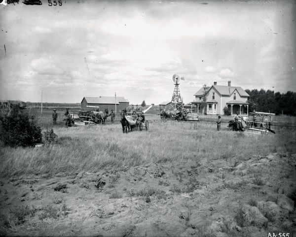 Group of men, children, women and horses posing with farm equipment in front of farm buildings, a farmhouse, and a windmill. A man is posing on a McCormick horse-drawn grain binder, and a man and a boy are posing in a horse-drawn carriage. There is a corn binder in the background. There are also two McCormick horse-drawn reapers — both have men sitting in the seats. Several other men are standing in the field near equipment. Women wearing bonnets are standing near the windmill.