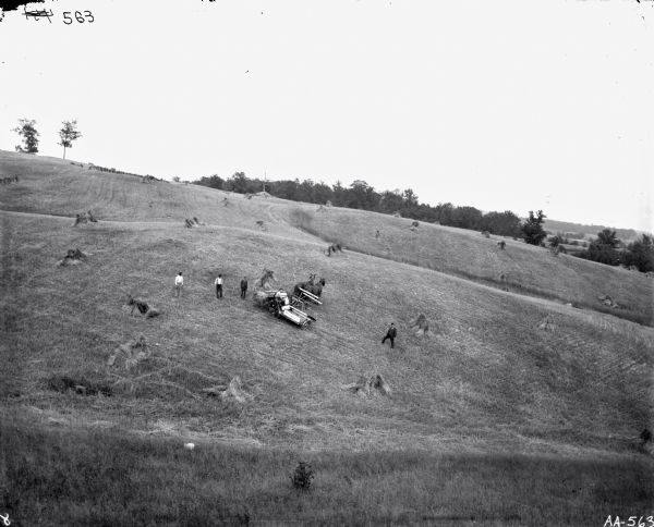 View from above of men posing on an opposite hill. One man is operating a horse-drawn McCormick grain binder, and four men (two wearing suits) are standing near him. There are several piles of grain in the field, and trees are in the background.