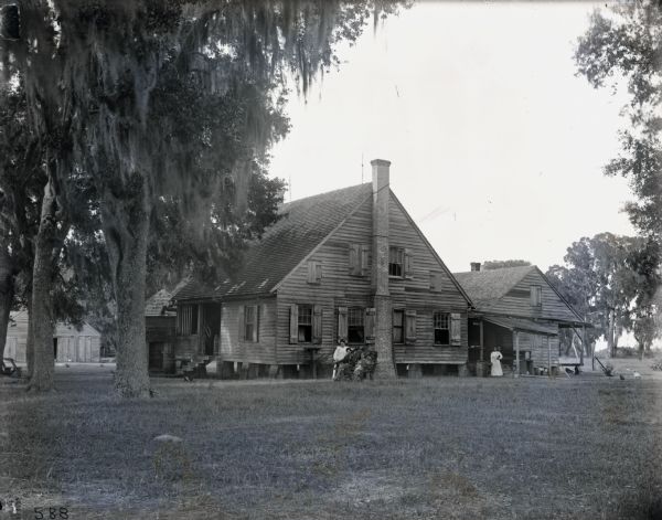 A boy and a girl are sitting on a bench in front of a house near Cypress trees. A woman wearing a white dress is standing behind them. The girl is holding a doll in her lap. On the far right an African American woman is standing alone near a smaller, separate dwelling. A barn is beside the house.