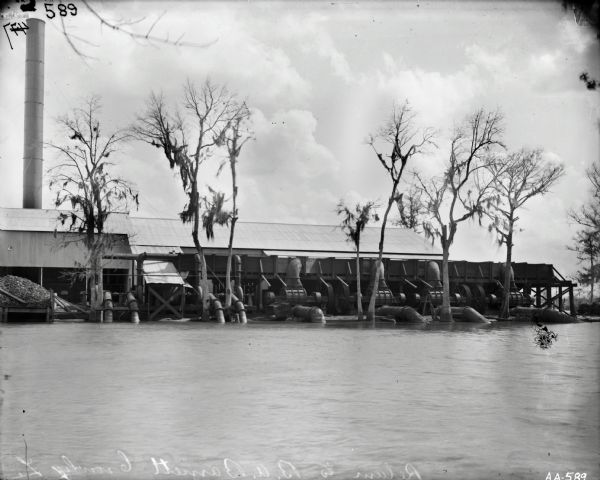 View across water of a factory or processing plant with large metal pipes leading into the water. Trees are growing out of the water near the pipes, and a smokestack rises above the building. The text scratched into glass plate reads: "Return to B. A. Barnett [Crowley] La". Possibly near or in Crowley, Louisiana.