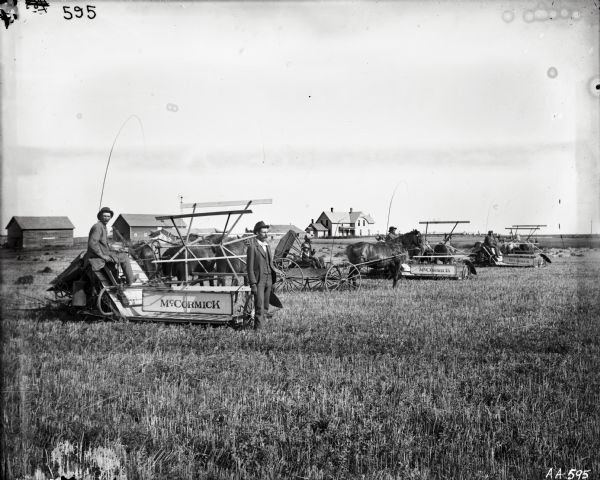 Men are posing in a row of horse-drawn McCormick grain binders. A man and child are posing in a horse-drawn carriage. In the background is a farmhouse and farm buildings.