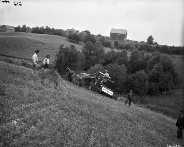 Four men are standing in a field on a steep hill near a man operating a horse-drawn McCormick grain binder. Two of the men are wearing suits. Farm buildings, a group of trees, and a fence are in the background.