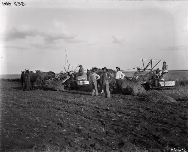 Group portrait of four men and a young girl posing in a field with two horse-drawn McCormick grain binders. Three of the men are standing with their hands on their hips. Behind them on the left a man is sitting in a horse-drawn grain binder, and on the right a young girl is sitting on another horse-drawn grain binder.