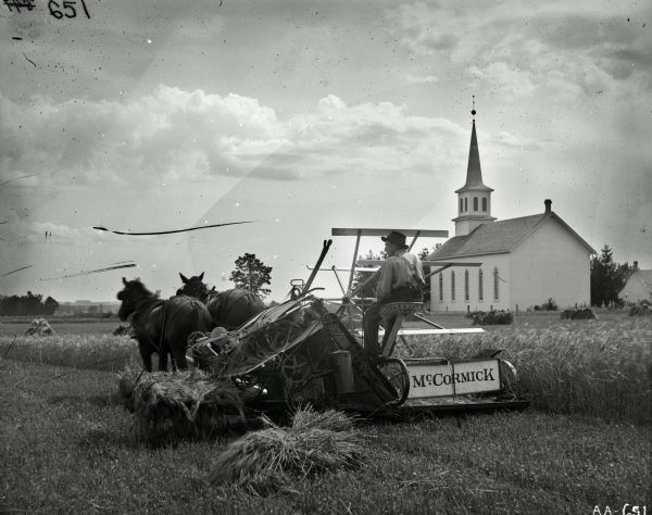 A man is operating a horse-drawn McCormick grain binder in a field of grain. A small pile of grain is beside him on the ground. A church and another building are in the background.