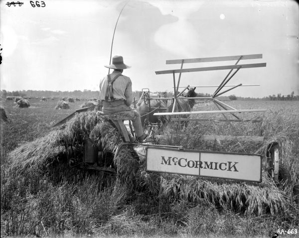 View from behind of a man operating a horse-drawn McCormick grain binder in a field. The man is wearing a large hat and holding a long whip. Piles of grain are distributed throughout the field. A line of trees is in the far background.