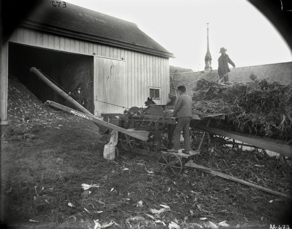 Two men are operating a McCormick husker-shredder. One man is feeding corn from a pile into the machine with a pitchfork. The shredded husks are being shot into a barn while the corn is bagged. Another man is standing inside the barn.
