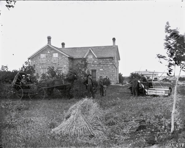 Men are posing around a horse-drawn grain binder while two women are looking on from a horse-drawn carriage. One of the men is holding a bicycle, and a farm house is in the background.