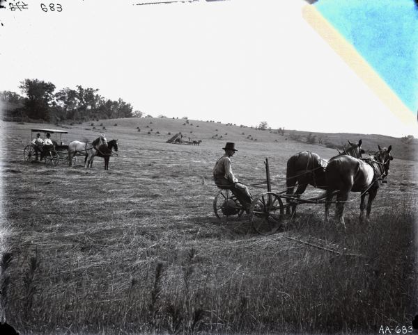 A man is sitting on a horse-drawn mower in a field, while two other men are looking on from a horse-drawn carriage.