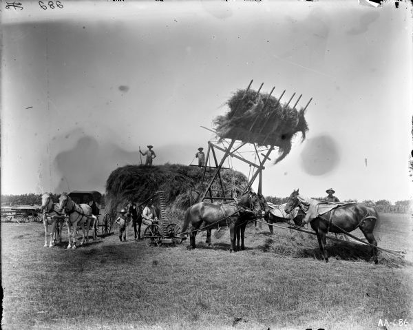Men are working with a horse-drawn mower and a horse-powered hay stacker in a field. Two men and a child are standing on a large stack of hay, and a horse-drawn carriage is parked nearby.