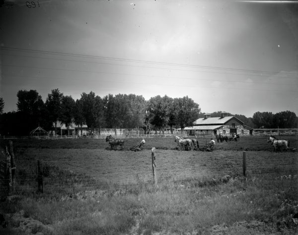 View over fence towards men using horse-drawn mowers in a field, while a couple in a horse-drawn carriage are looking on. Fences and farm buildings are in the background.