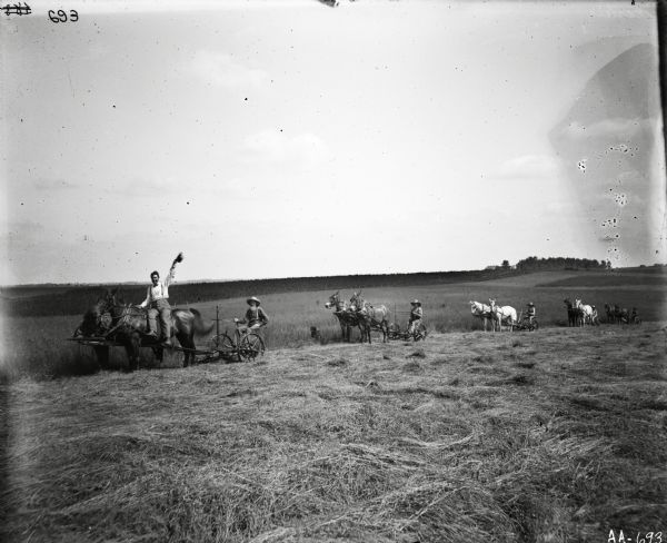 A line of men and horse-drawn mowers in a field. One man wearing a bow tie is sitting astride a horse and waving his hat at the camera.