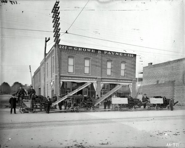 Men and boys are posing with McCormick husker-shredders (corn shredders) in the street in front of Crowe and Payne, a McCormick dealership. Signs over plate glass windows read: "farm implements" and "carriages & wagons."