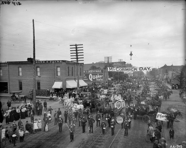 Elevated view of people, horses, and farm machines gathered on the town's main street for a "McCormick Day" celebration, with several grain binders and a brass band. "McCormick Days" were organized by McCormick dealers to celebrate the delivery of new machines to customers. Dealers used the events to promote the company and generate further sales. This event was likely organized by McCormick dealers Crowe and Payne.
