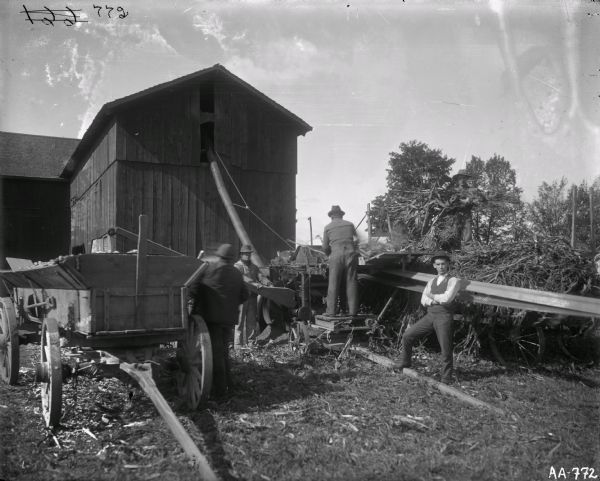 Four men are working with a husker-shredder and a wagon near a barn. The husker-shredder is powered with a belt hooked to what is probably a tractor (not visible).