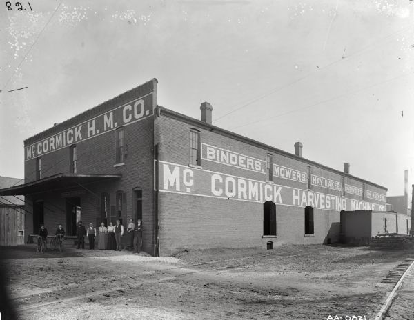 View across railroad tracks of men and women standing in front of a McCormick Harvester Machine Company dealership or branch house on a dirt road in an unidentified town. Two young men are sitting on bicycles nearby, and railroad box cars are parked along the side of the building.