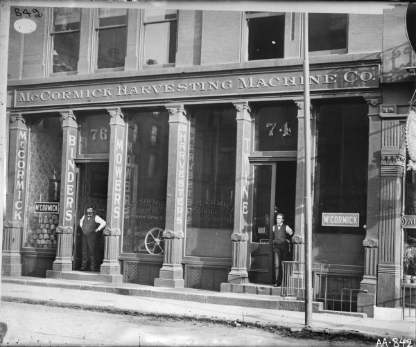 View across street of dealership or agency building of the McCormick Harvesting Machine Company. Two men stand outside the storefront at two entrances. There are signs advertising mowers, binders, harvesters and twine. The man on the left may be the general agent, Frank Culbertson.