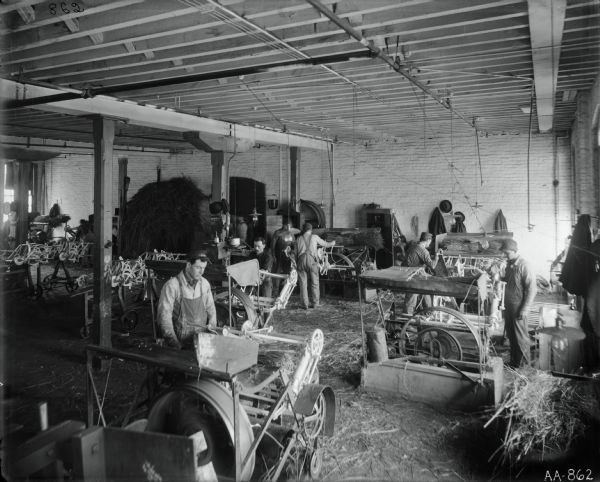 Slightly elevated view of factory workers testing(?) the binding mechanisms for grain binders at the McCormick Reaper Works. Loose hay and hay bundles are distributed around the factory floor.