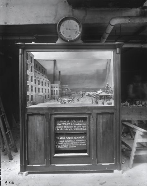 A wooden exhibit cabinet, apparently recently constructed, on what appears to be a factory or shop floor with a work table, ladder, saw horse and wood shavings. The case includes a diorama with factory buildings, rural houses and a man with a horse-drawn grain binder. Below the diorama, text in English and French reads: "A Host of Machines. Over 2,000,000 McCormick machines are in use throughout the world, equal to the labor in harvest of 20,000,000 men." There is also a dial or clock at the top of the case with notches from one to sixty. A man is faintly visible in the background at right. The case may have been constructed for the McCormick exhibit at the 1900 Paris Exposition.