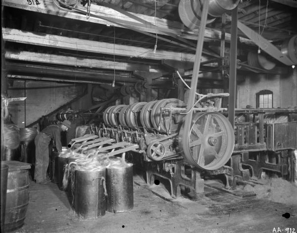 A male employee of the McCormick Twine Mill works with sisal fiber as it comes out of a finisher machine and piles into metal buckets. The machine is belt-driven from the ceiling.