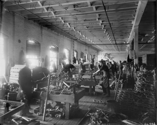 Factory workers assemble grain binder parts at the McCormick Reaper Works. Metal machinery pieces are stacked at right and men work at tables to the left near a row of windows.
