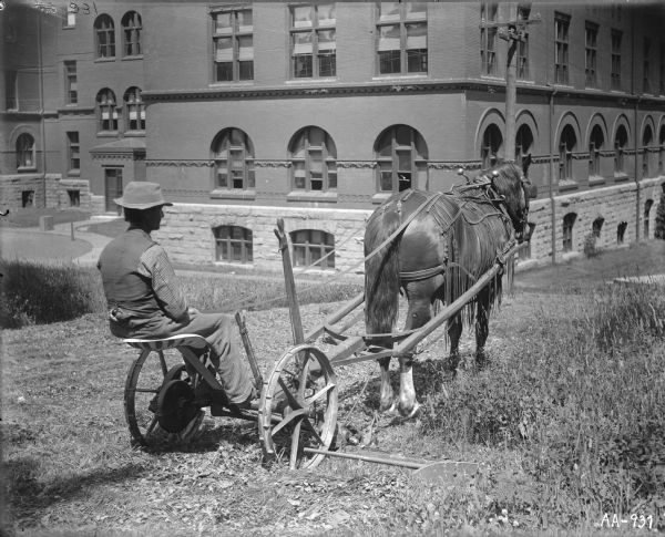 Man using a horse-drawn mower on the grounds of a large institutional building. The horse is wearing a fly-net.