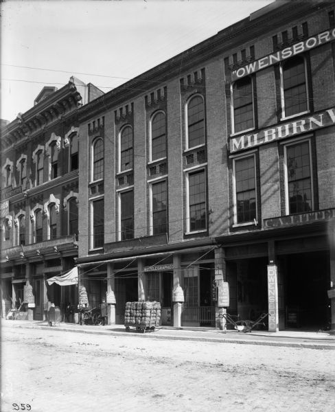 Storefronts along a city street, including a McCormick Harvesting Machine Company dealership, and a Western Union Telegraph office. A cart loaded with bundles is parked on the sidewalk near a plow. A sign nearby reads "The Owensboro." A small group of men, including a police officer is standing to the left on the sidewalk.