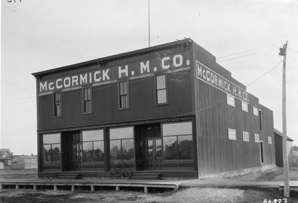 Exterior view of McCormick Harvesting Machine Company dealership building. Large windows on ground floor of window reflect surrounding landscape. A raised boardwalk or sidewalk runs in front of the building, and a bicycle is parked on it.