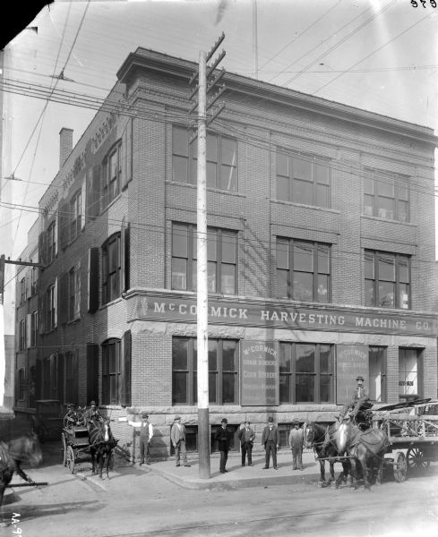 View across street of storefront of a McCormick Harvesting Machine Company dealership. Men are standing along the sidewalk. Two other men are sitting on horse-drawn wagons holding the reins of their horses.