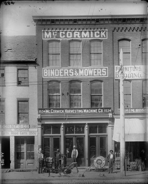 Elevated view from across street of men posing with farm and lawn equipment along the storefront of a McCormick Harvesting Machine Company dealership. Three African American employees(?) and another man (possibly the General Agent, W.K. Bache) are standing with a horse-drawn mower, bundles of twine, and push mowers.