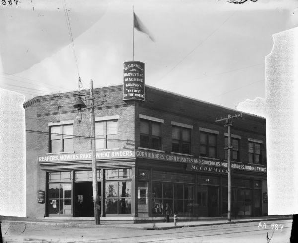 Exterior of a McCormick Harvesting Machine Company dealership or general agency building. A sign on the roof of the building reads "the best in the world." A man is standing on the sidewalk in front of the building.