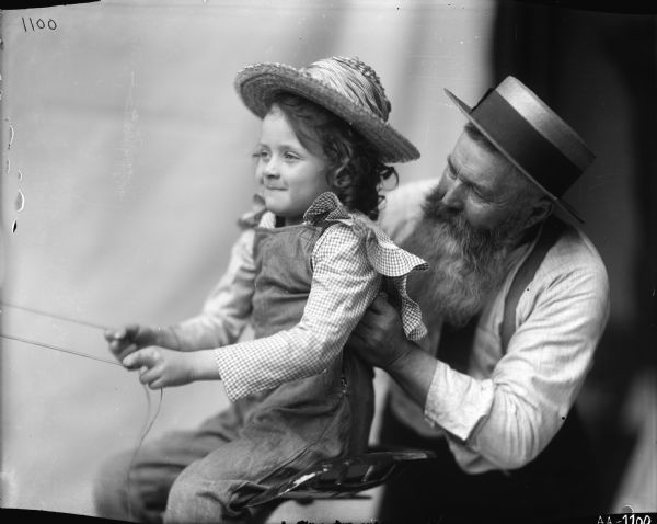 A young girl is sitting on the seat of a farm implement and holding onto reins as an older bearded man is assisting her. The image was likely used as a model for advertising art.