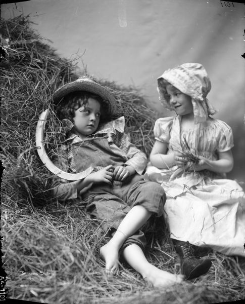 A boy and girl gaze at one another in a haystack, one with a reaping hook and the other with a handful of straw. The children were models and the photograph was used as the basis of a McCormick advertising poster.